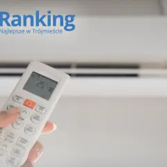 Dłoń trzymająca pilot do klimatyzacji na tle białego urządzenia.