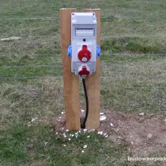 Tymczasowe gniazda elektryczne na budowie, zasilane prądem budowlanym, zamontowane na drewnianym słupku przy ogrodzeniu.