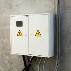 Biała skrzynka elektryczna z żółtymi symbolami ostrzegawczymi na betonowej ścianie.