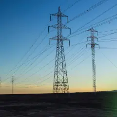 Wieże energetyczne niosące prąd przemienny na tle zachodzącego słońca.