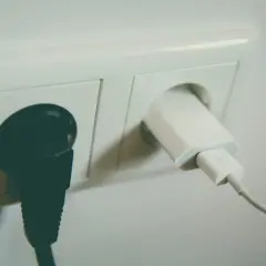 Dwa gniazdka elektryczne. W jednym czarna wtyczka, w drugim biała ładowarka z kablem. Napięcie w gniazdku zasila urządzenia.