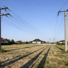 Dwa betonowe słupy wysokiego napięcia przecinają niebo nad pustą działką. W tle widać domy mieszkalne.