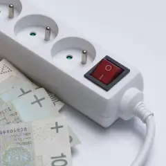 Biała listwa zasilająca z czerwonym włącznikiem i polskimi banknotami. Taki dodatek do prądu może oznaczać wysokie rachunki.