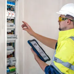 Elektryk w kasku i kamizelce odblaskowej sprawdza rozdzielnicę elektryczną na tablecie.