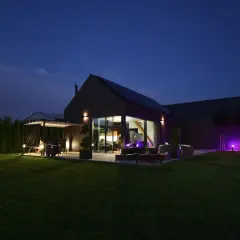 Nowoczesny dom pasywny oświetlony nocą. Widać taras z grillem, duże okna, ogród i fioletowe lampy.