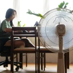 Biały wentylator stojący na pierwszym planie, w tle kobieta przy biurku.