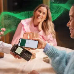 Kobieta płaci telefonem w restauracji. W tle uśmiechnięta przyjaciółka. Pozytywne fortum opinie o płatnościach zbliżeniowych.