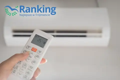 Dłoń trzymająca pilot do klimatyzacji na tle białego urządzenia.