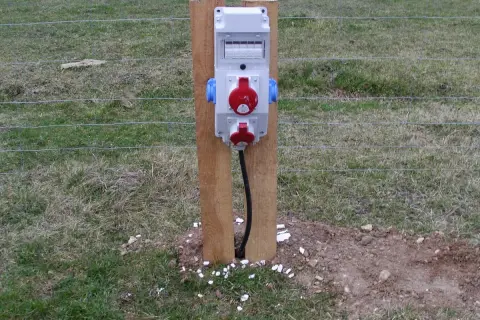 Tymczasowe gniazda elektryczne na budowie, zasilane prądem budowlanym, zamontowane na drewnianym słupku przy ogrodzeniu.