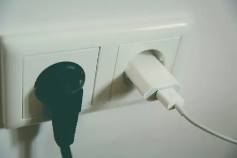 Dwa gniazdka elektryczne. W jednym czarna wtyczka, w drugim biała ładowarka z kablem. Napięcie w gniazdku zasila urządzenia.