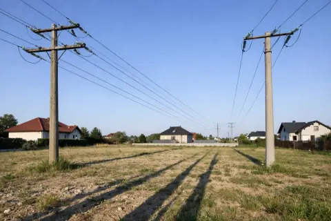Dwa betonowe słupy wysokiego napięcia przecinają niebo nad pustą działką. W tle widać domy mieszkalne.
