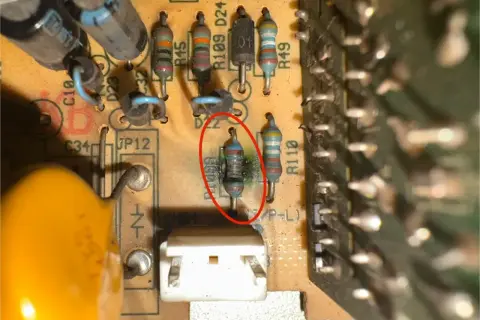 Uszkodzony rezystor z czerwonym okręgiem, otoczony innymi elementami elektronicznymi na płytce drukowanej.