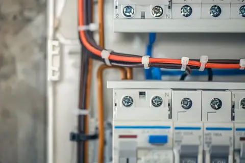 Wnętrze rozdzielnicy elektrycznej z przewodami w kolorach pomarańczowym i brązowym, zabezpieczonymi opaskami. Widoczne wyłączniki i zaciski.