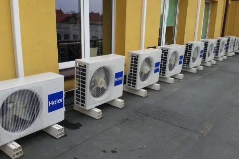 Rząd białych jednostek klimatyzacyjnych Haier na dachu, obok żółtej ściany budynku.