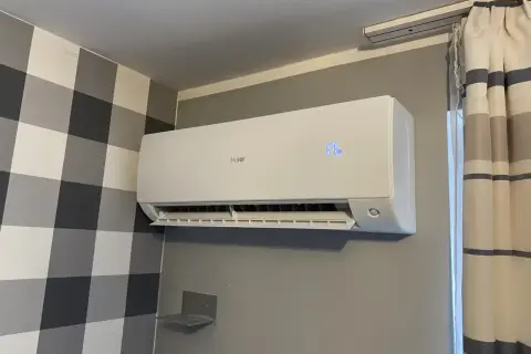 Biała klimatyzacja Haier zamontowana na szarej ścianie, obok wzorzystej tapety i zasłon.