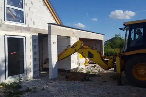 Żółta koparko-ładowarka pracuje przy budowie domu z bloczków. Widać okna i drzwi. Trwa wlz.