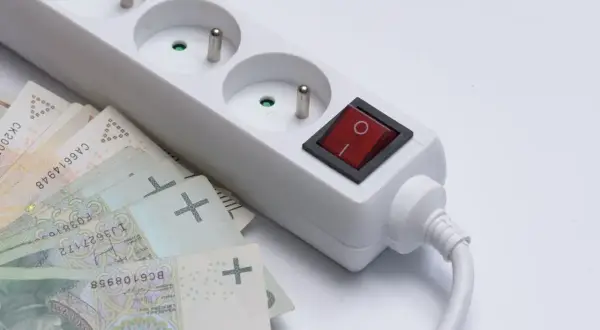 Biała listwa zasilająca z czerwonym włącznikiem i polskimi banknotami. Taki dodatek do prądu może oznaczać wysokie rachunki.