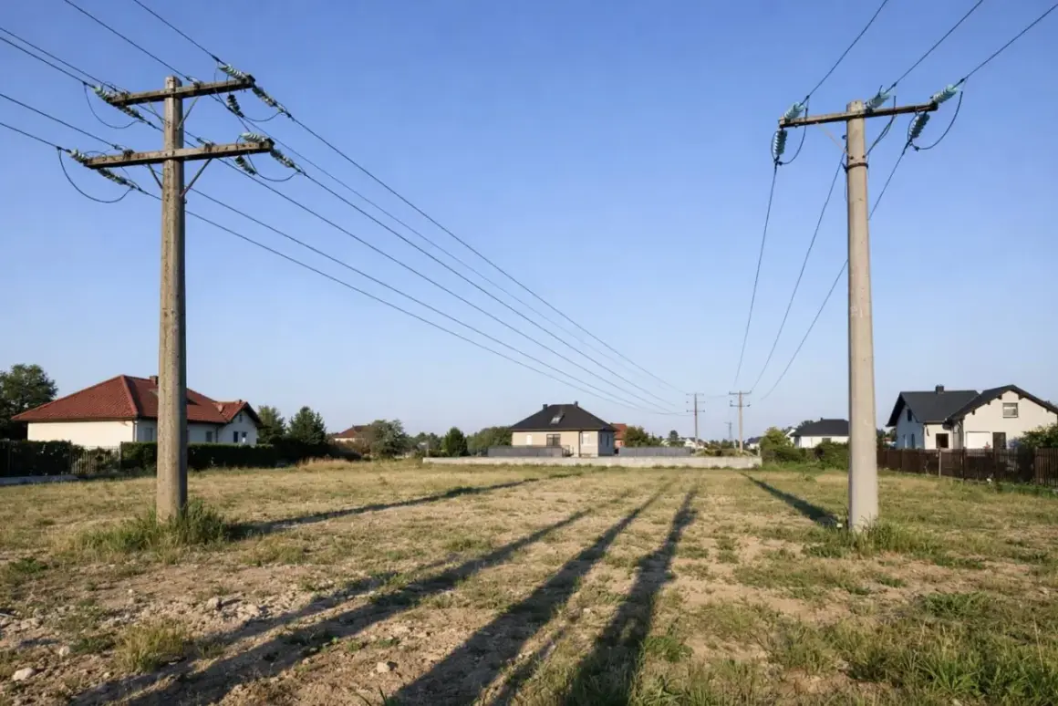 Dwa betonowe słupy wysokiego napięcia przecinają niebo nad pustą działką. W tle widać domy mieszkalne.