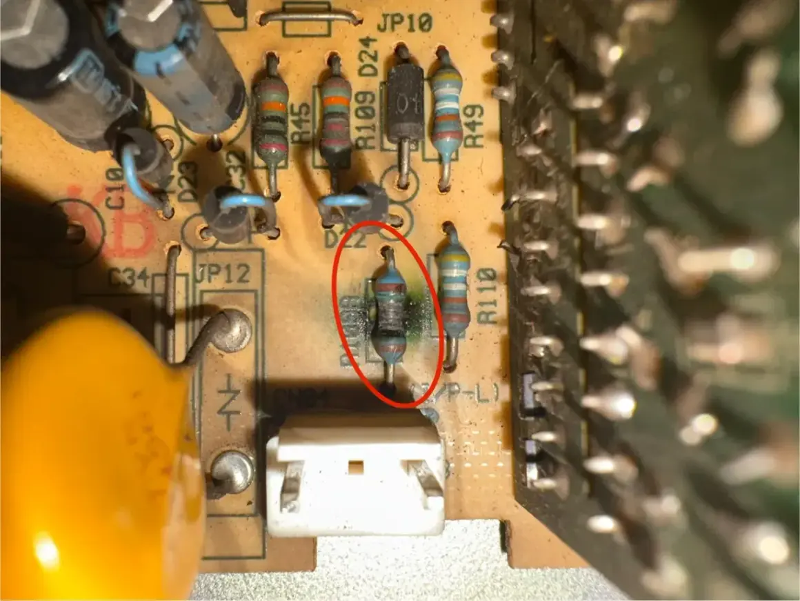 Uszkodzony rezystor z czerwonym okręgiem, otoczony innymi elementami elektronicznymi na płytce drukowanej.