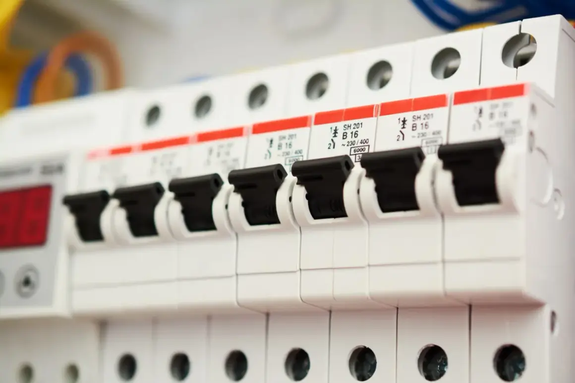 Szereg białych wyłączników automatycznych, każdy z czarną dźwignią. Na czerwonych etykietach widnieją oznaczenia, sugerujące instalację prądu trójfazowego.