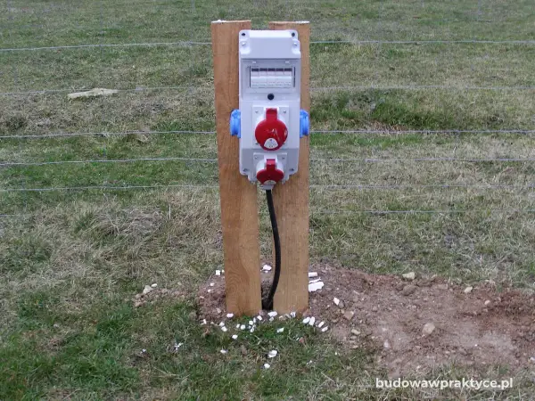 Tymczasowe gniazda elektryczne na budowie, zasilane prądem budowlanym, zamontowane na drewnianym słupku przy ogrodzeniu.