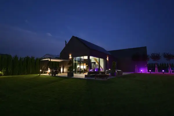 Nowoczesny dom pasywny oświetlony nocą. Widać taras z grillem, duże okna, ogród i fioletowe lampy.