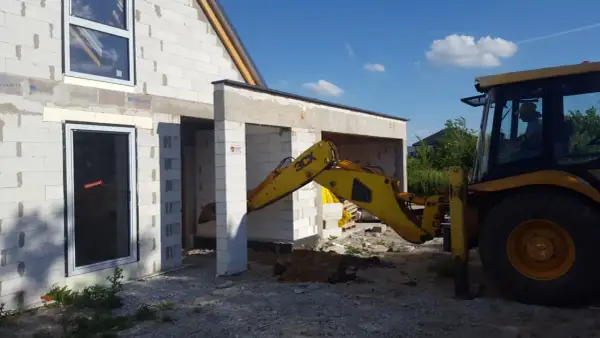Żółta koparko-ładowarka pracuje przy budowie domu z bloczków. Widać okna i drzwi. Trwa wlz.