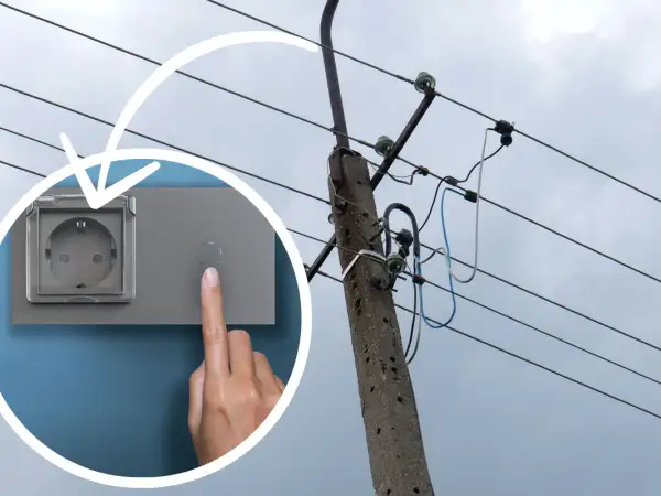 Ręka naciska przycisk obok gniazdka, sugerując koszt instalacji elektrycznej. W tle słup energetyczny z przewodami.
