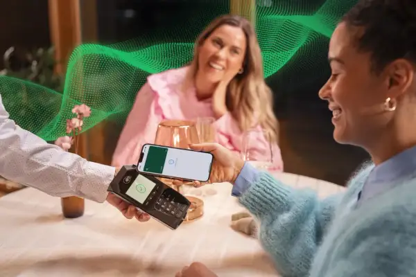 Kobieta płaci telefonem w restauracji. W tle uśmiechnięta przyjaciółka. Pozytywne fortum opinie o płatnościach zbliżeniowych.